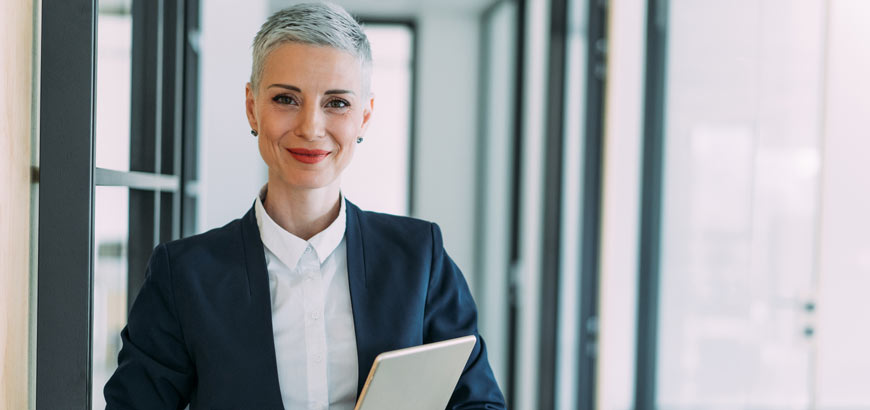 A woman with short gray hair in a suit holding a tablet, standing in a modern office hallway.