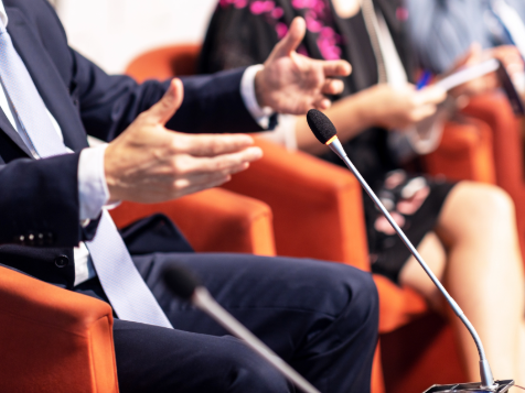 People sit in orange chairs with microphones, one person gesturing with hands while speaking.