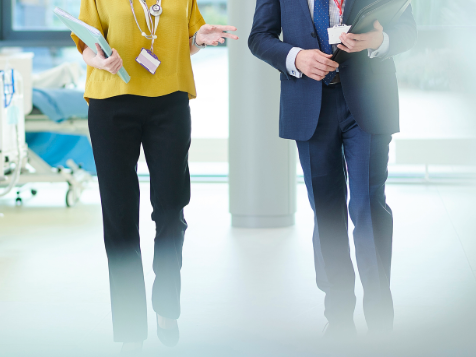Two professionals walk through a hospital corridor holding folders and wearing ID badges.