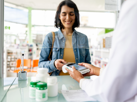A person in a denim jacket pays at a pharmacy counter using a card reader held by a pharmacist, with medication bottles on the counter.
