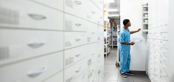 Person in blue scrubs stands in a narrow aisle between white cabinets, reaching into an open cabinet on the right.