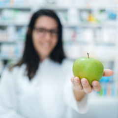 Person in a white coat holding a green apple, with shelves of products blurred in the background.