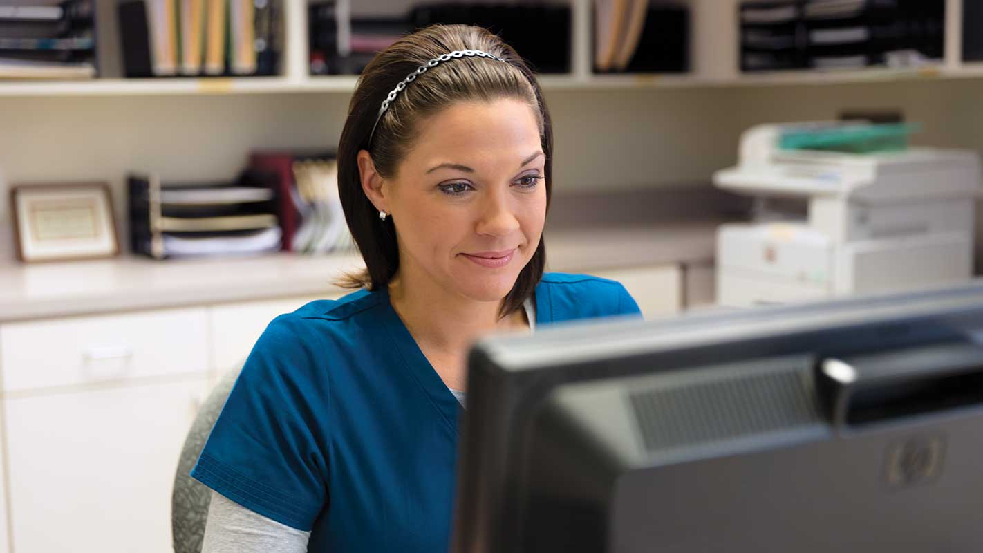 Person in blue scrubs sitting at a desk with a computer monitor, office shelves in the background.