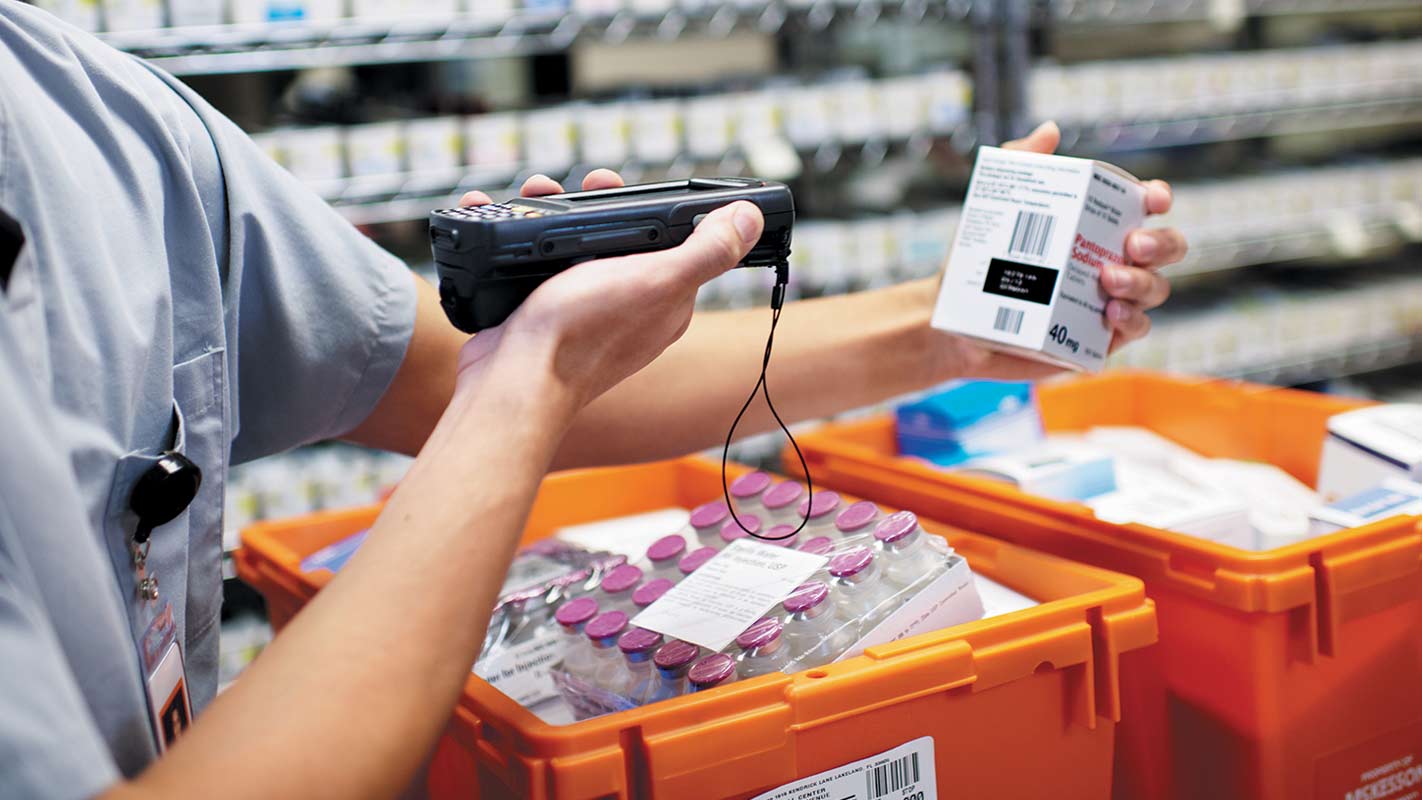 A person scans a medication box in a pharmacy with an orange bin of medications in the foreground.