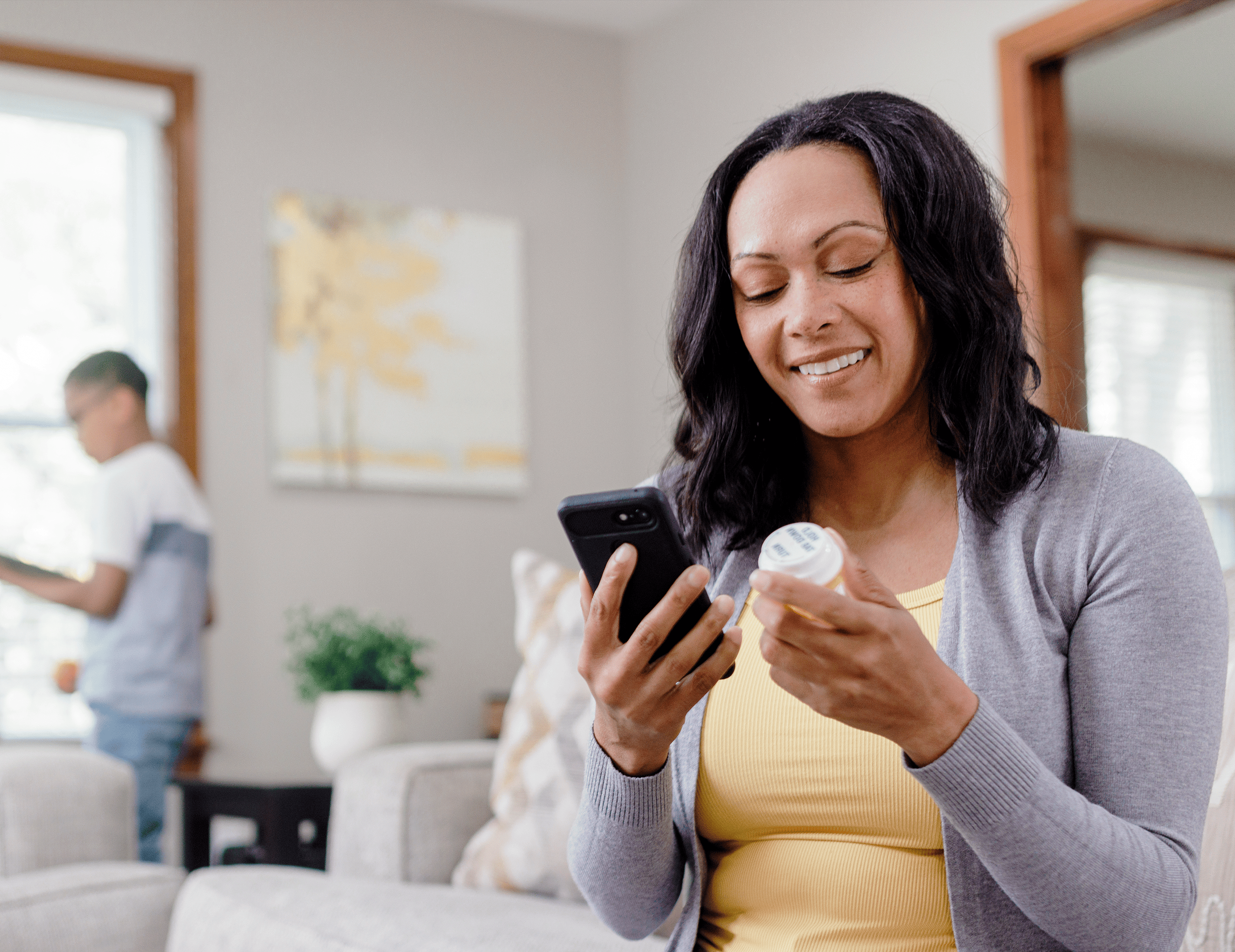 A person in a yellow shirt and gray cardigan holds a smartphone and a pill bottle, with a child in the background.