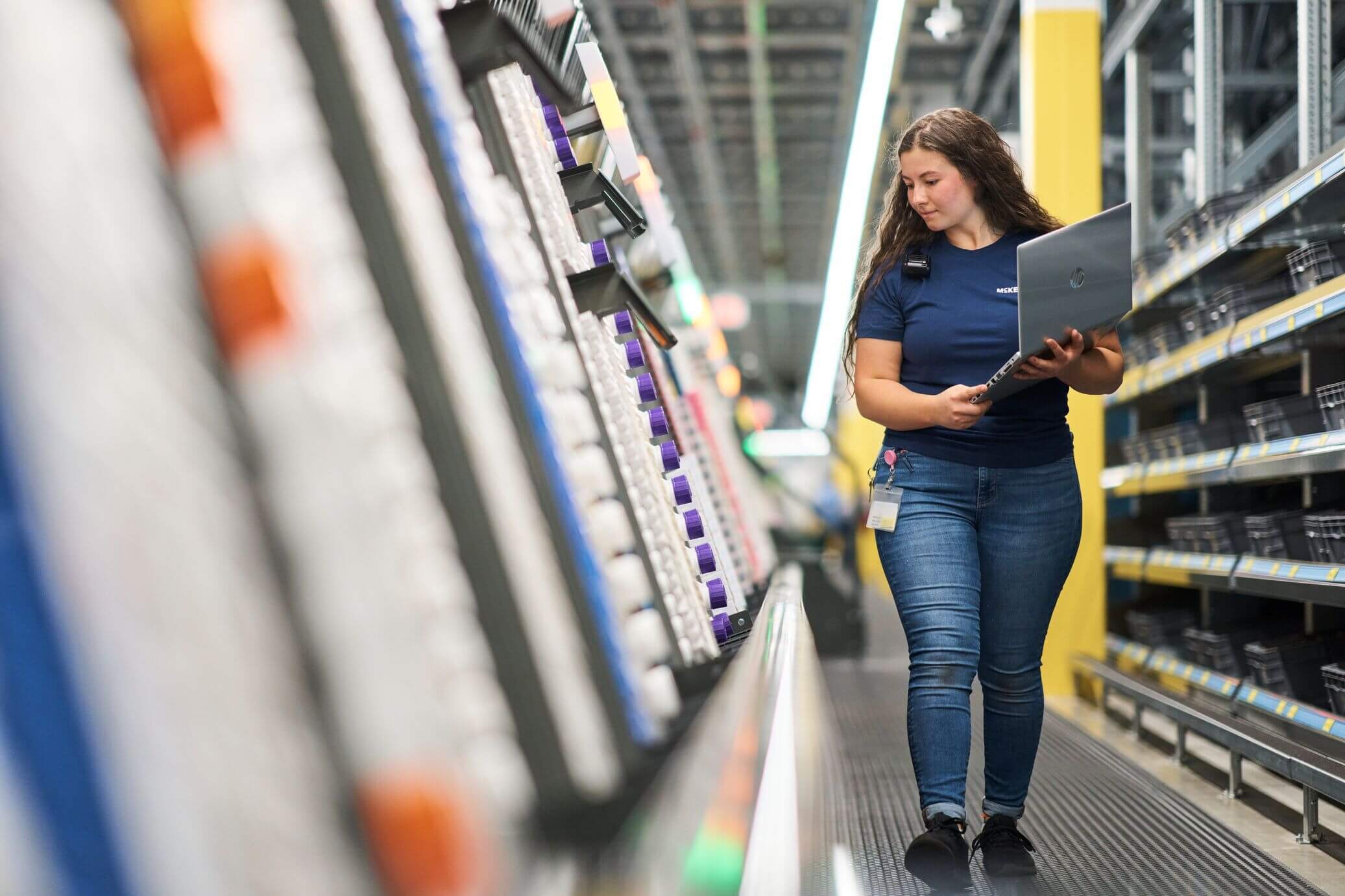 Person in a blue shirt and jeans holding a laptop, walking through an aisle in a warehouse or store with shelves of products on both sides.