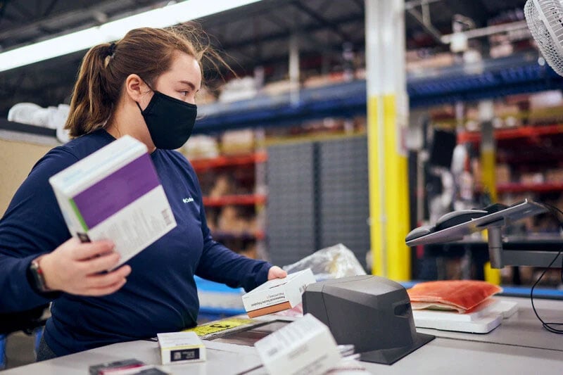 A person in a warehouse setting is holding several books while standing at a workstation with a scanner and other equipment.