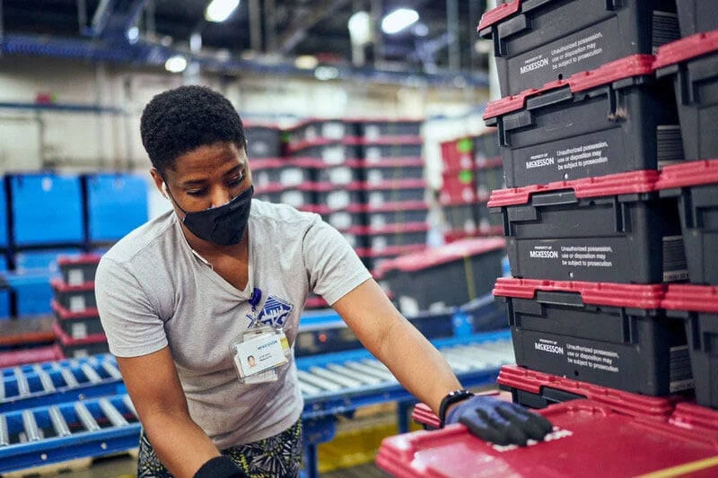 A person in a warehouse stacks red and black plastic bins labeled "McKesson" on a conveyor belt. They wear a name badge and gloves.