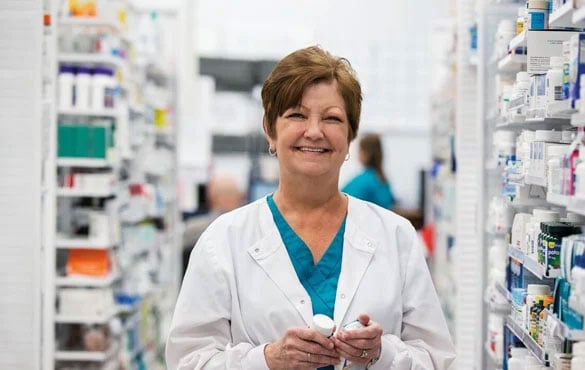 Pharmacist in a white coat holding a medication bottle, standing between shelves of pharmaceutical products in a pharmacy.