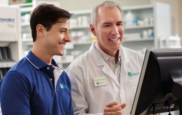 Two pharmacy staff members, one in a blue polo shirt and the other in a white lab coat, are looking at a computer screen in a pharmacy setting.
