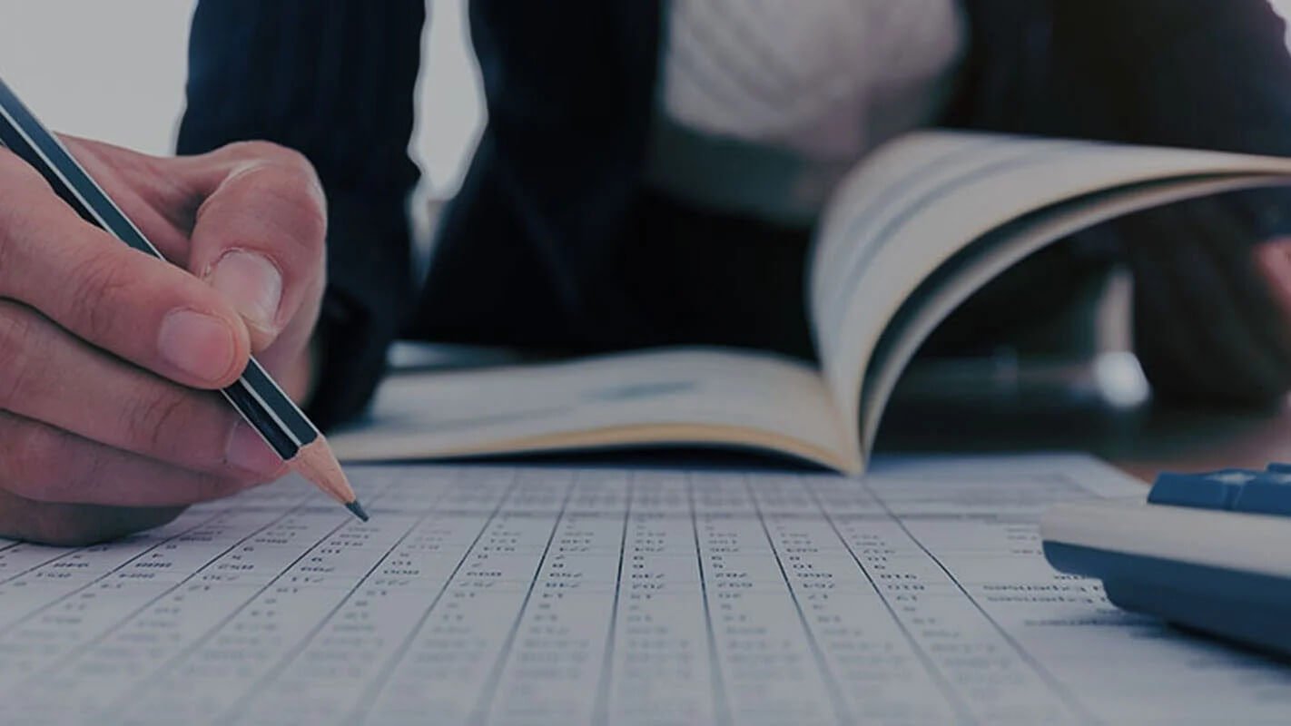 Person writing with a pencil on a spreadsheet, with an open book and calculator nearby.