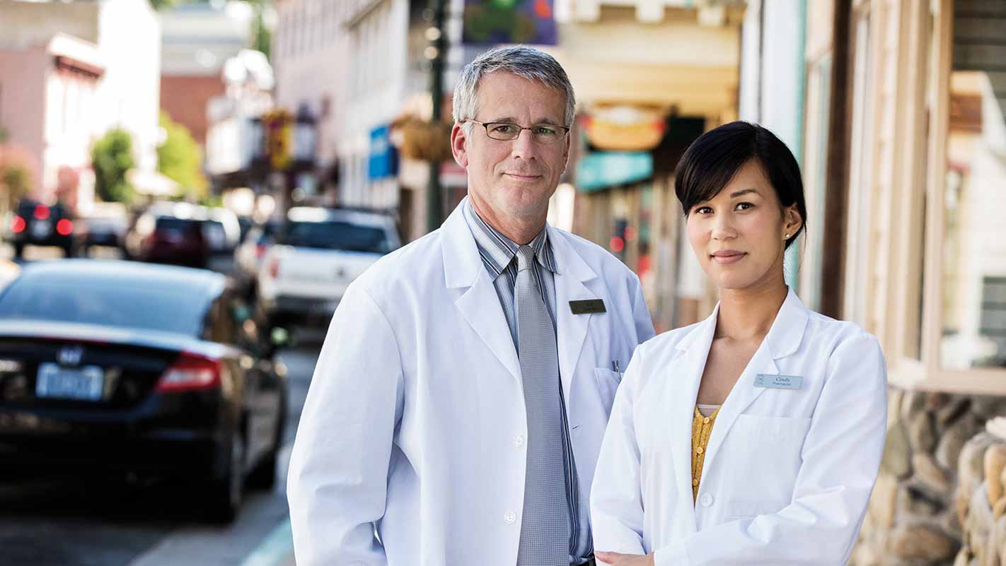 Two individuals wearing white lab coats stand on a street. The background shows parked cars and buildings.