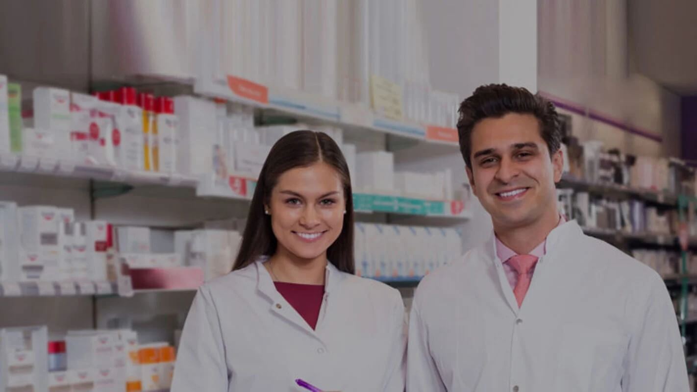 Two people in white lab coats standing in a pharmacy aisle with shelves of various medical products behind them.