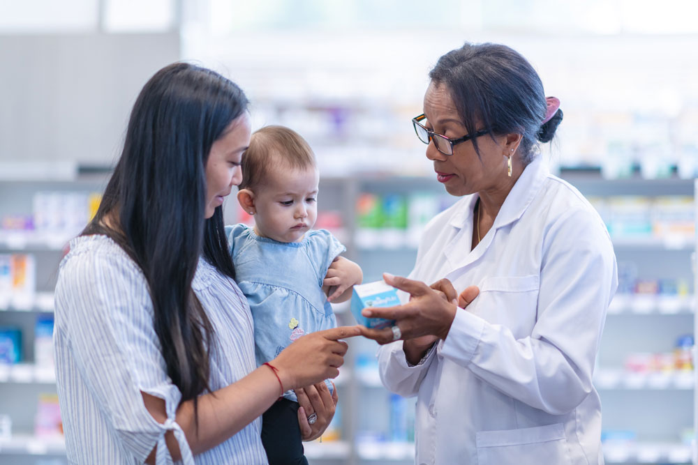 Pharmacist explains medication to a woman holding a baby inside a pharmacy.