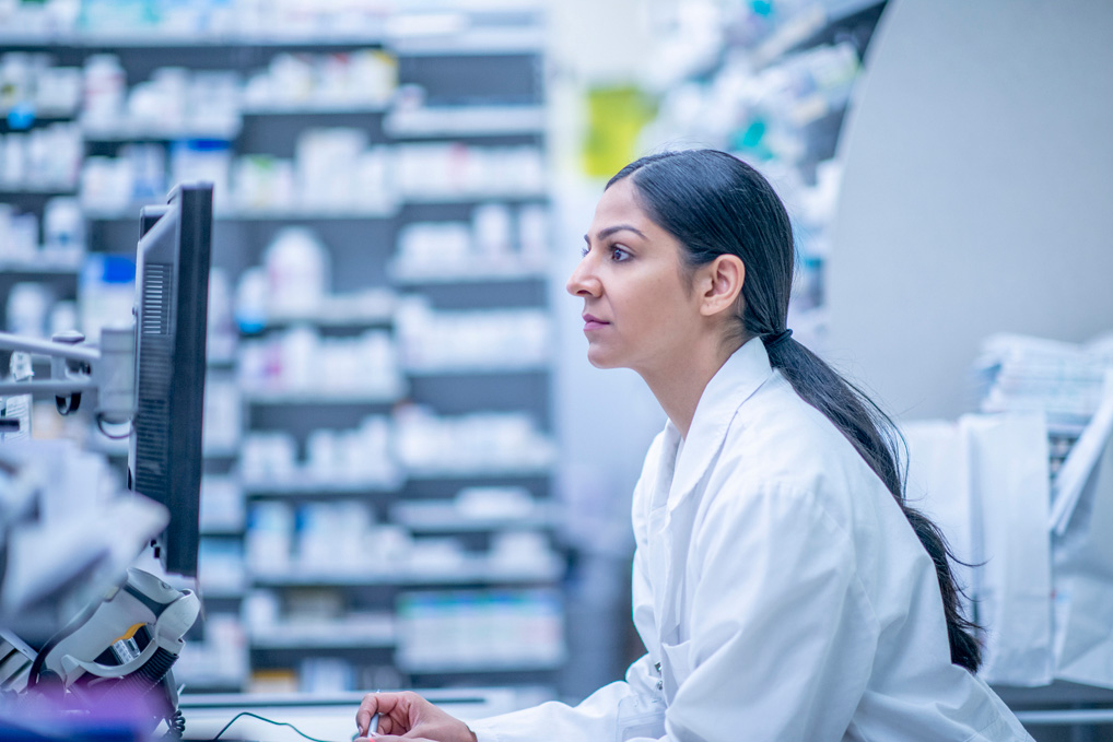 Pharmacist in a white coat working at a computer in a pharmacy with shelves of medications.