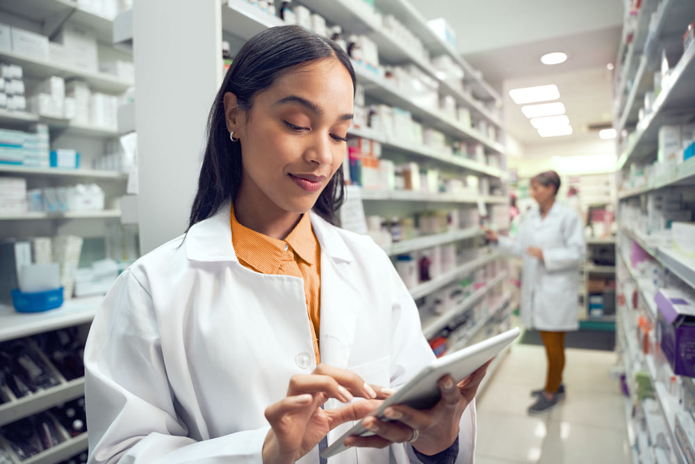 Two pharmacists in white coats work in a pharmacy, one with a tablet and one organizing shelves.