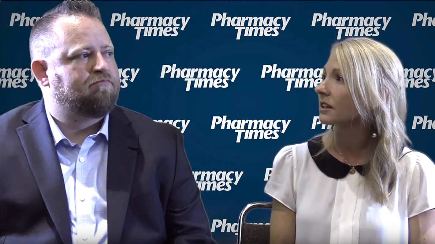 Two people sit in front of a backdrop that reads "Pharmacy Times." One wears a suit, the other a white blouse with a black collar.
