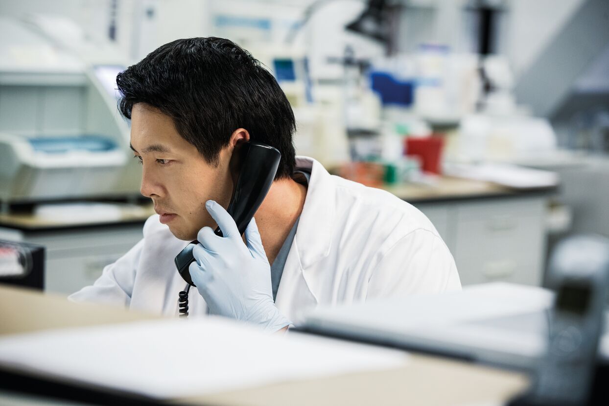 A person in a lab coat and gloves is talking on the phone in a laboratory setting.