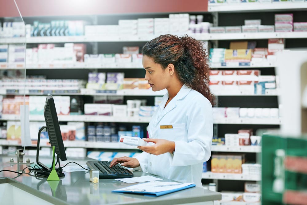 Pharmacist in a white coat holds medication while typing on a computer in a pharmacy.