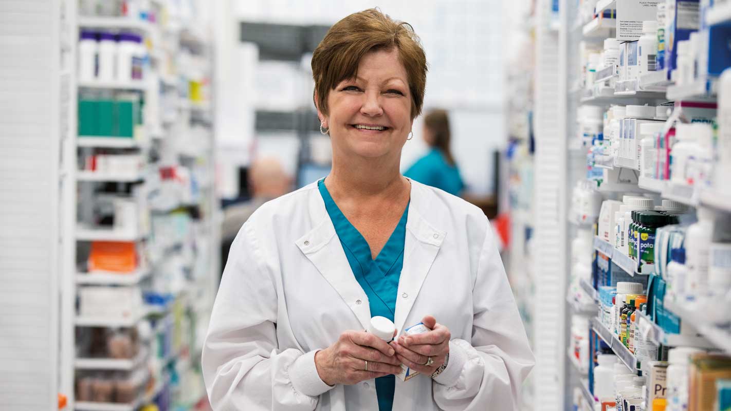 Pharmacist holding a medication bottle in a pharmacy aisle filled with various medications.