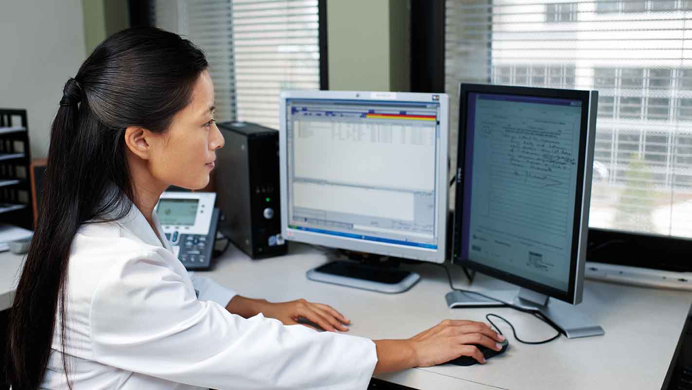 A person in a white coat working on a computer with dual monitors in an office setting.
