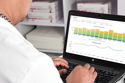 A person in a white coat uses a laptop displaying bar and line graphs, with shelves of medical supplies in the background.