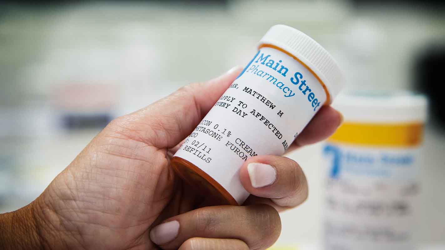 Close-up of a hand holding a prescription bottle labeled "Main Street Pharmacy" with instructions for use.