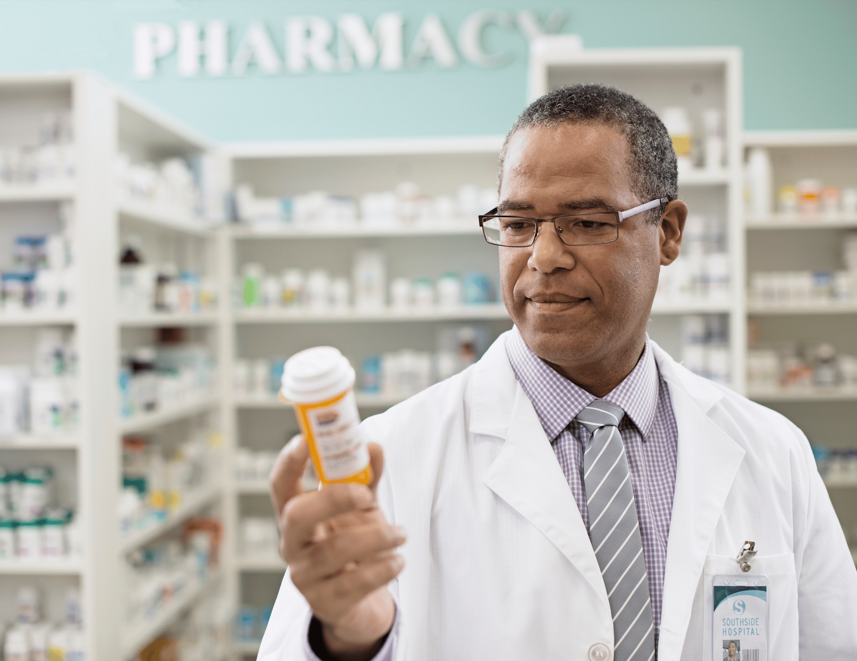 Pharmacist in a white coat holding a prescription bottle in a pharmacy, with shelves of medications and the word "PHARMACY" visible in the background.