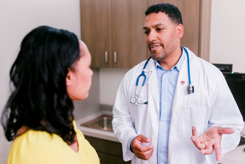 A doctor wearing a white coat and stethoscope talks to a patient in a yellow shirt in a medical office.