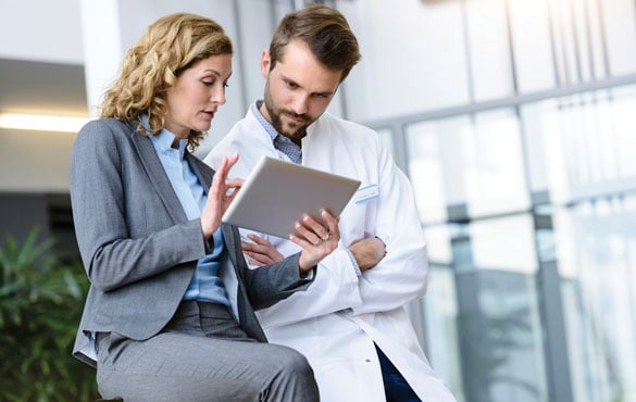 A person in a suit and a person in a lab coat look at a tablet together.