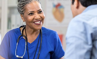 A healthcare professional in blue scrubs with a stethoscope talks to a person.