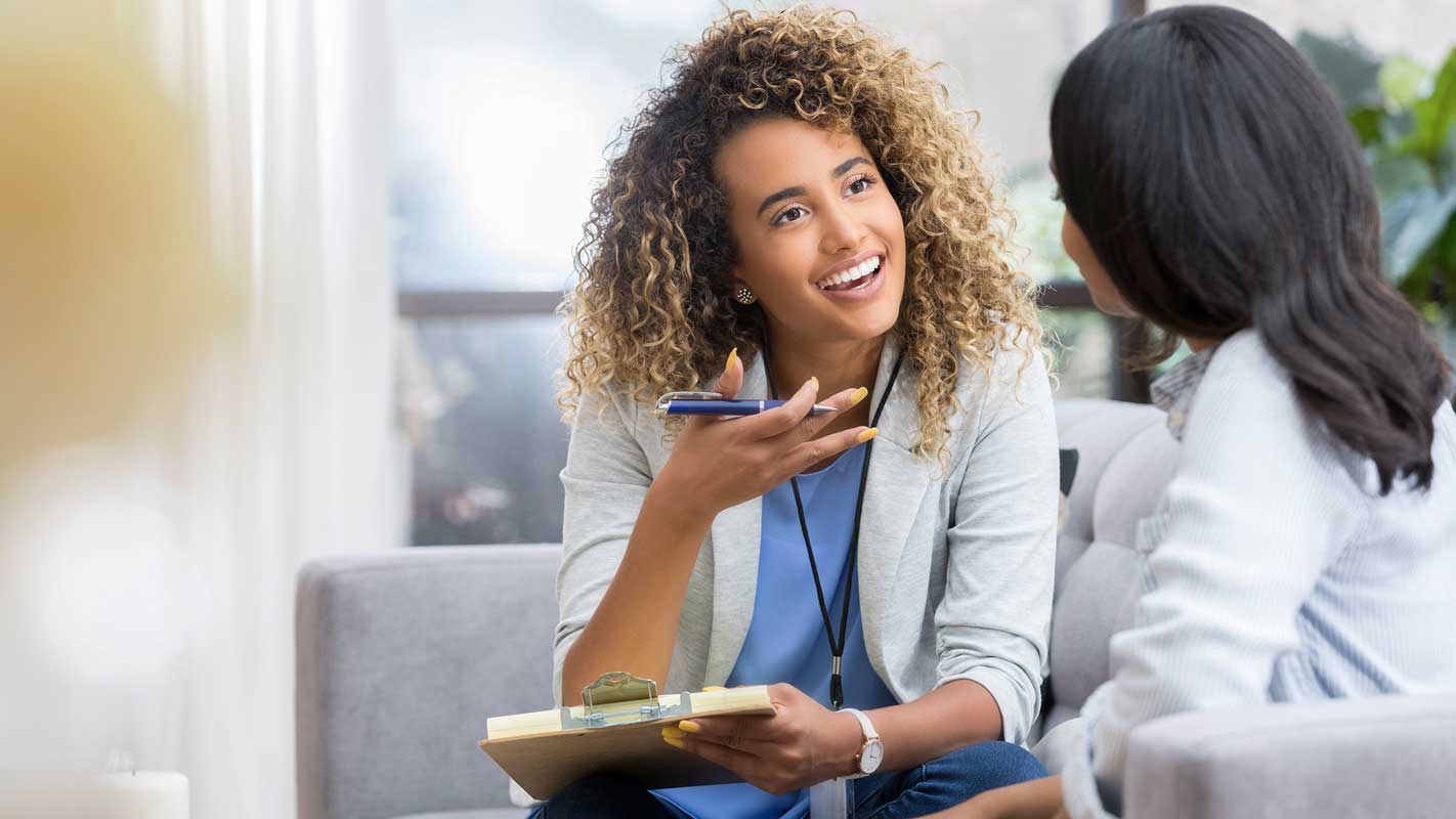 A person with curly hair holding a pen and clipboard, talking to another person on a couch.