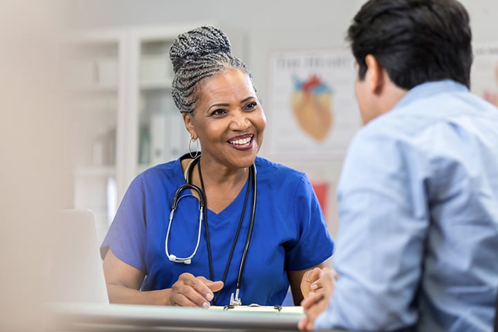 A healthcare professional in blue scrubs with a stethoscope speaks to a patient in an office with medical posters and shelves.