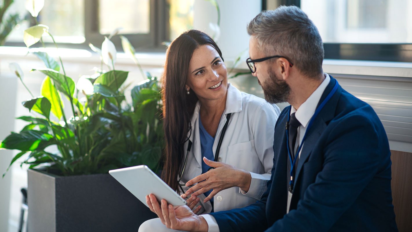 Two professionals, one in a white coat and one in a suit, discussing something on a tablet in an office with plants.
