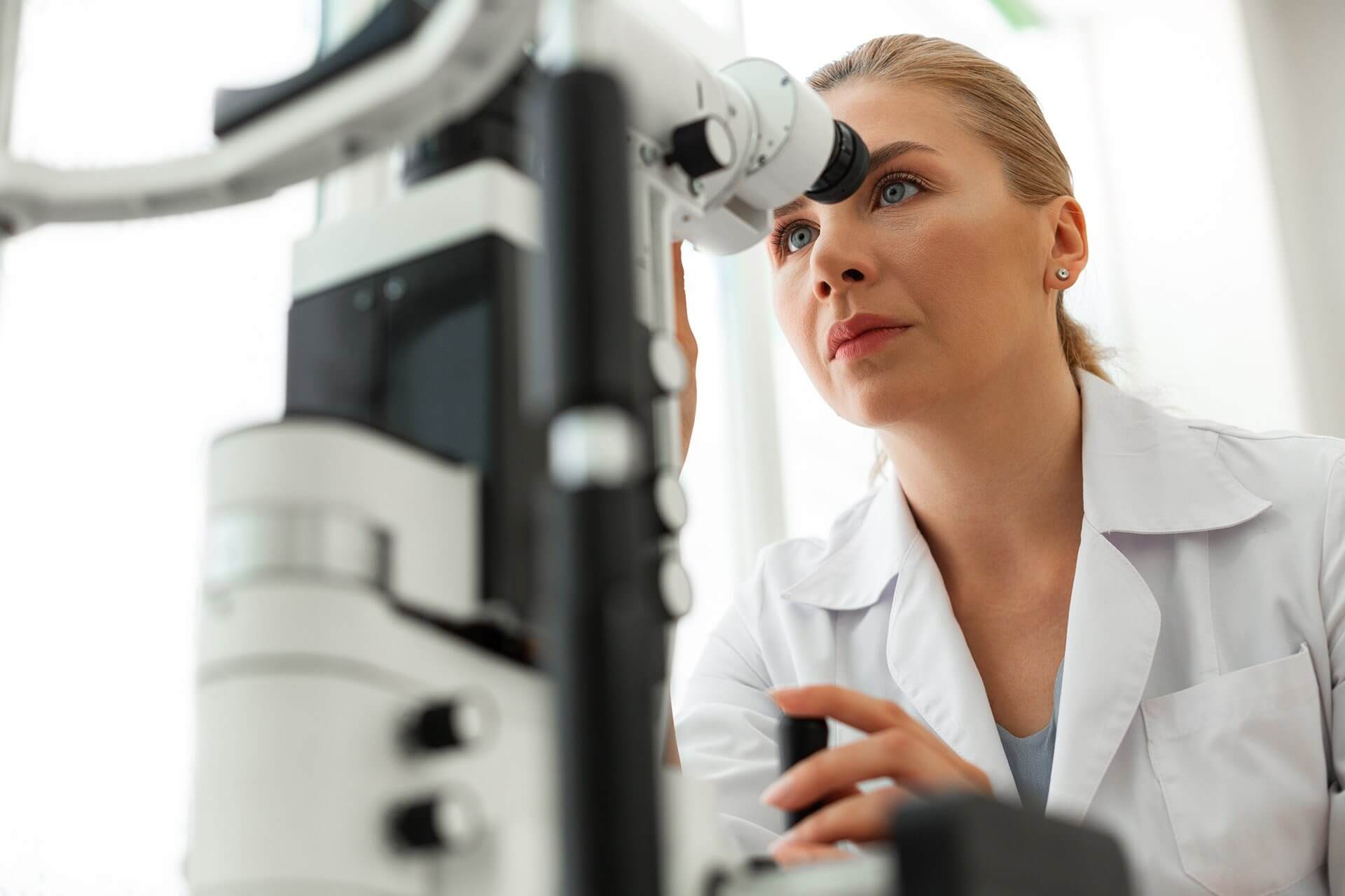 Person in a white lab coat using an optical medical device, face blurred.