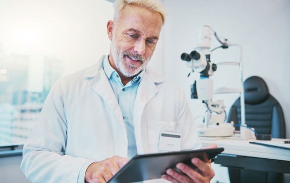 A doctor in a white coat uses a tablet in a medical office with an eye examination microscope in the background.