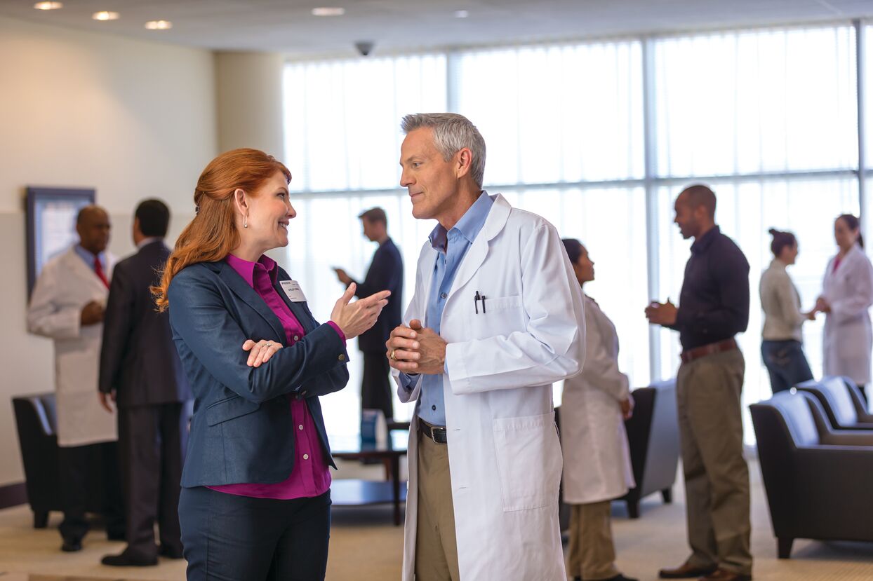 A group of medical professionals and business people are engaged in conversations in a hospital or clinic lobby. The setting is professional and collaborative.