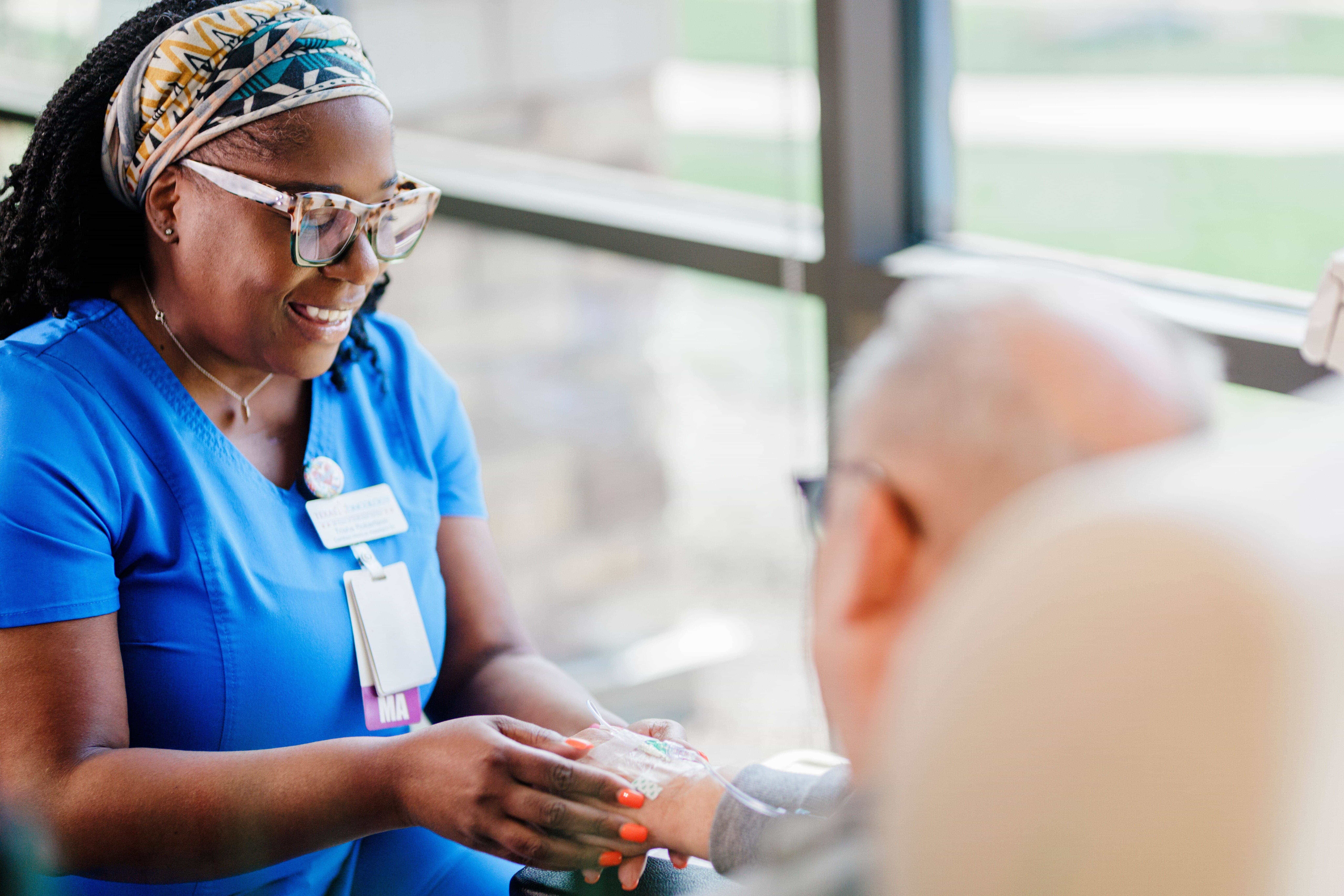 A healthcare worker in blue scrubs holds the hand of an elderly person in a medical setting.