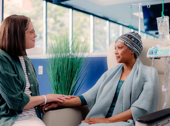 A person in a headscarf and hospital gown sits in a chair, holding hands with another person.