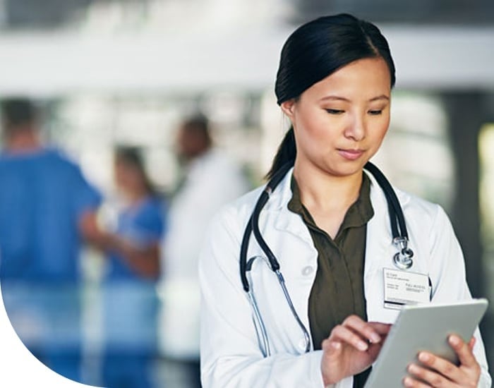 A doctor in a white coat uses a tablet, with blurred medical staff in the background.