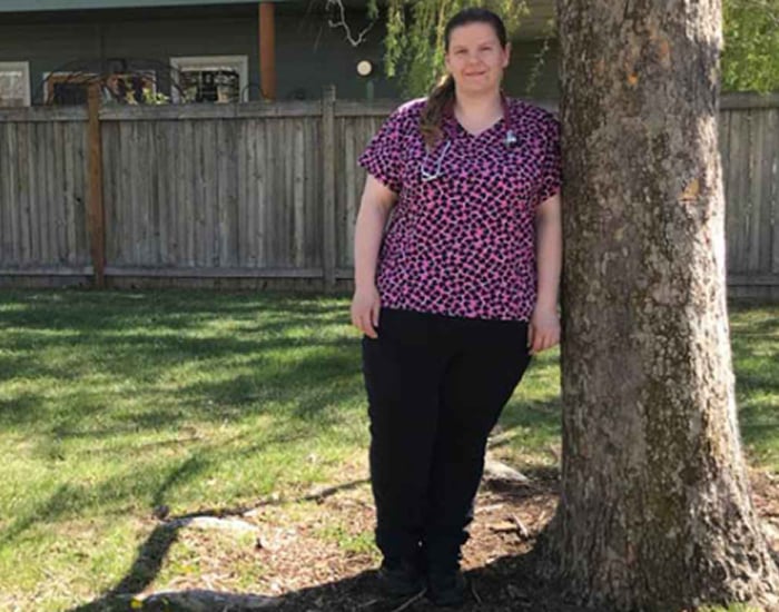 A person in a pink and black patterned shirt stands next to a tree in a grassy yard.