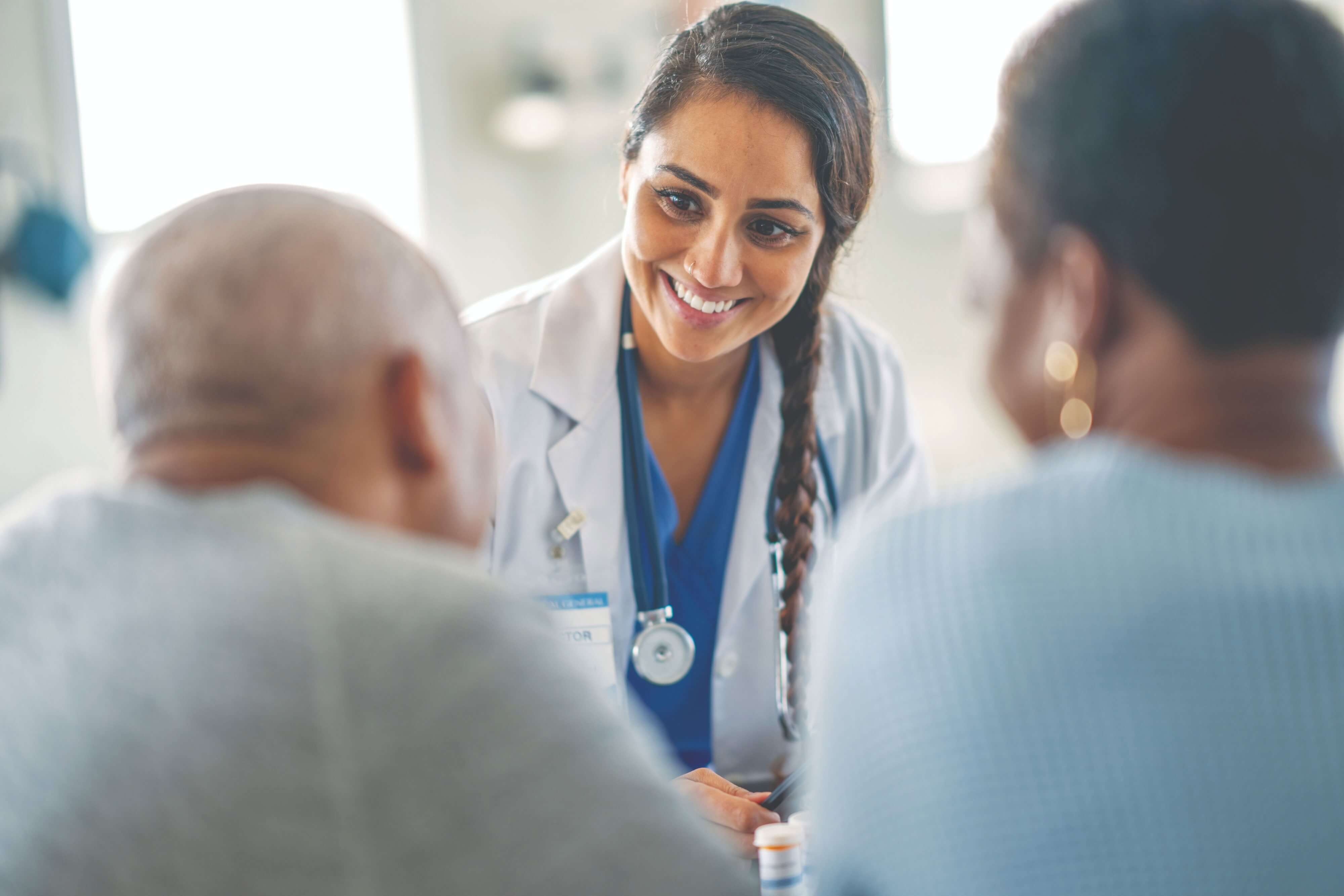 A doctor in a white coat speaks to two patients in a medical consultation room.