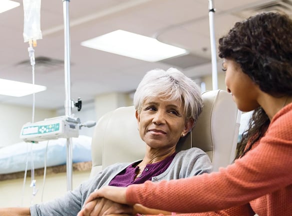 Two people in a medical setting, one receiving an IV drip and the other offering support.