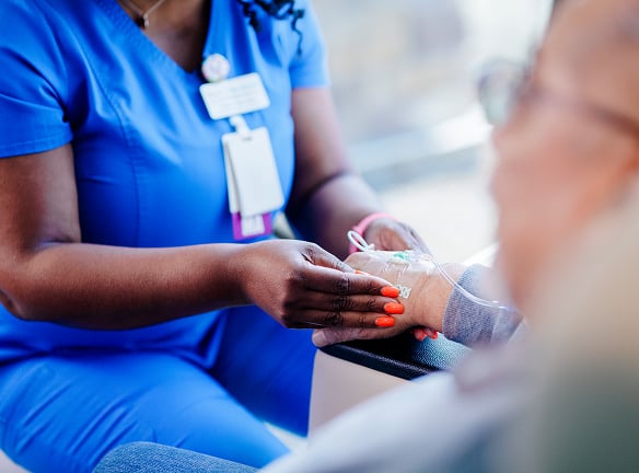 A nurse in blue scrubs adjusts an IV line on a patient's hand.