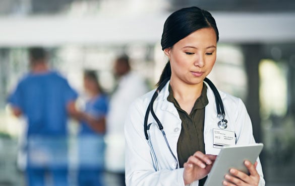 Medical professional in a white coat with a stethoscope around the neck, using a tablet. Blurred background shows other medical staff in blue scrubs.