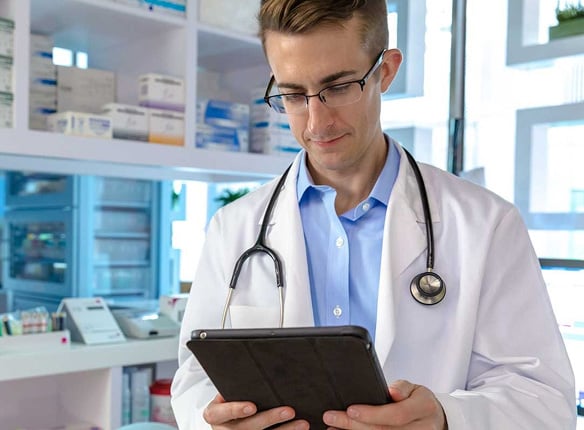 A doctor in a white coat with a stethoscope around the neck holds a tablet in a medical office.