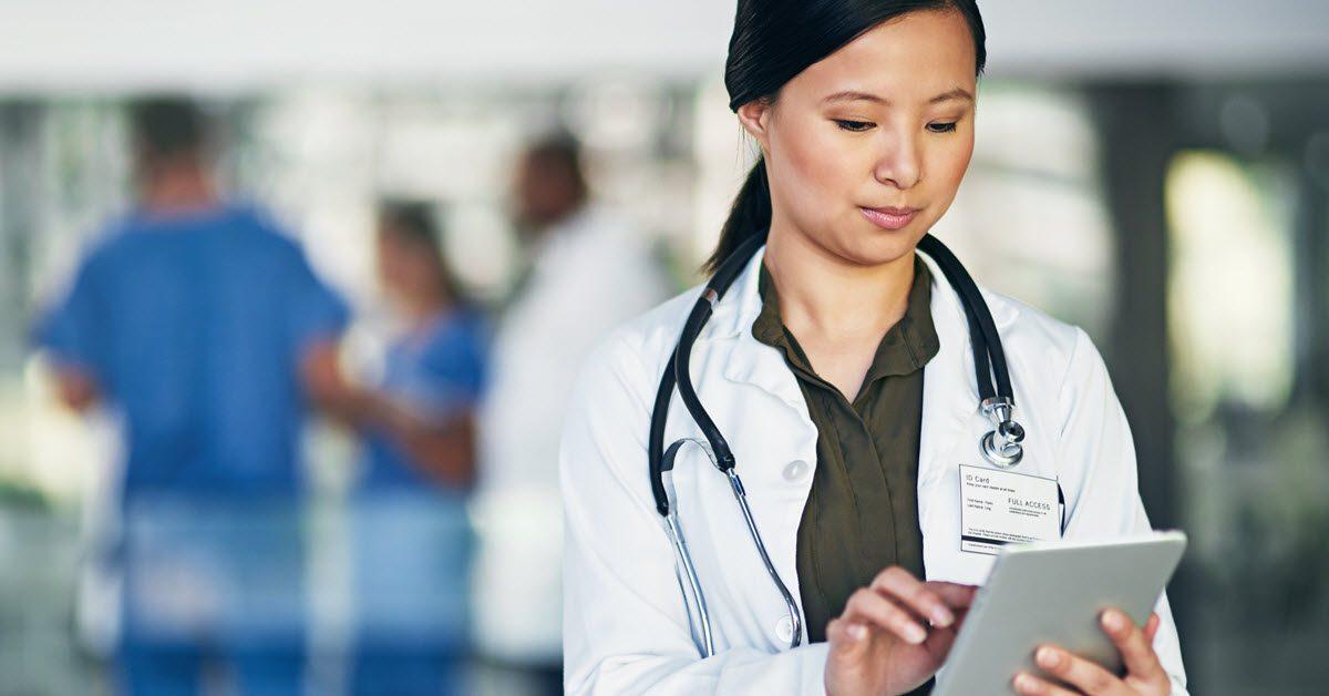 A doctor in a white coat uses a tablet, with blurred medical staff in the background.