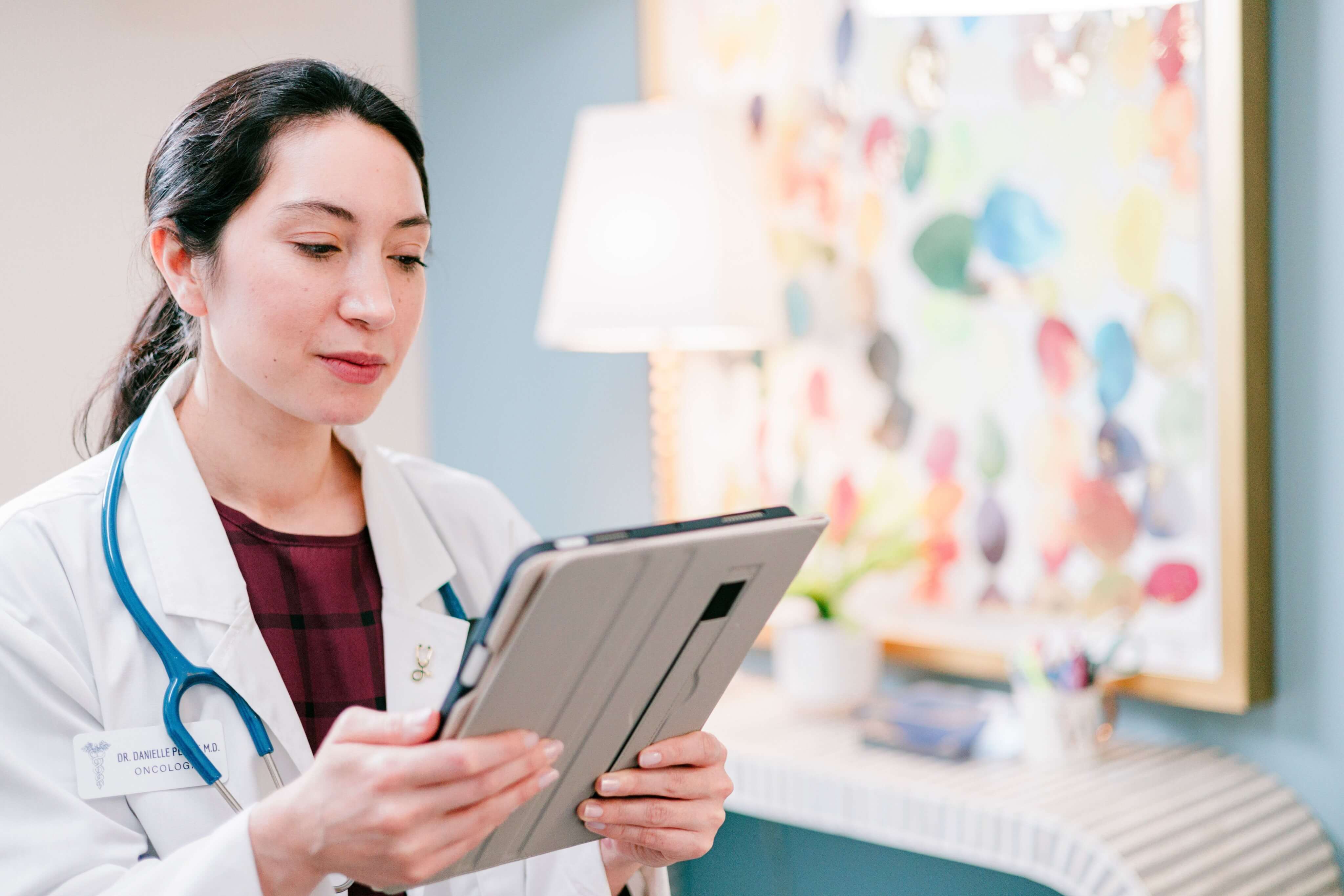 A doctor in a white coat holding a tablet, with a stethoscope around the neck, standing in an office with colorful artwork and a lamp in the background.