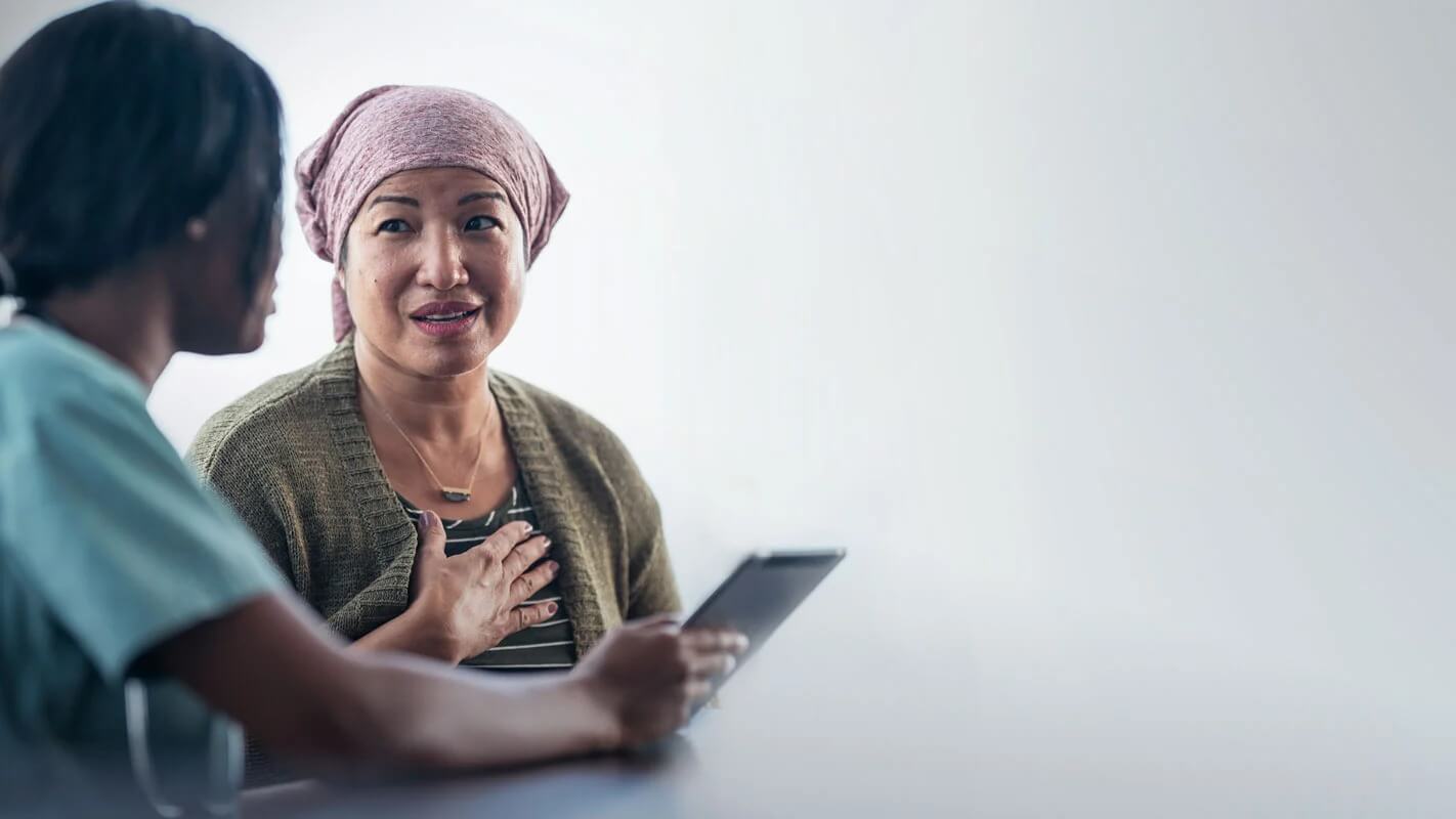 A healthcare professional in scrubs speaks with a patient wearing a headscarf, who holds a tablet and touches their chest.