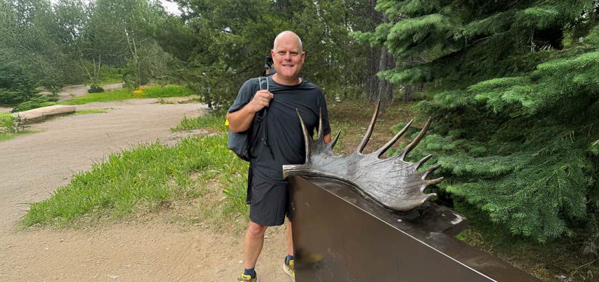 A person stands beside a large moose antler sculpture in a wooded park.